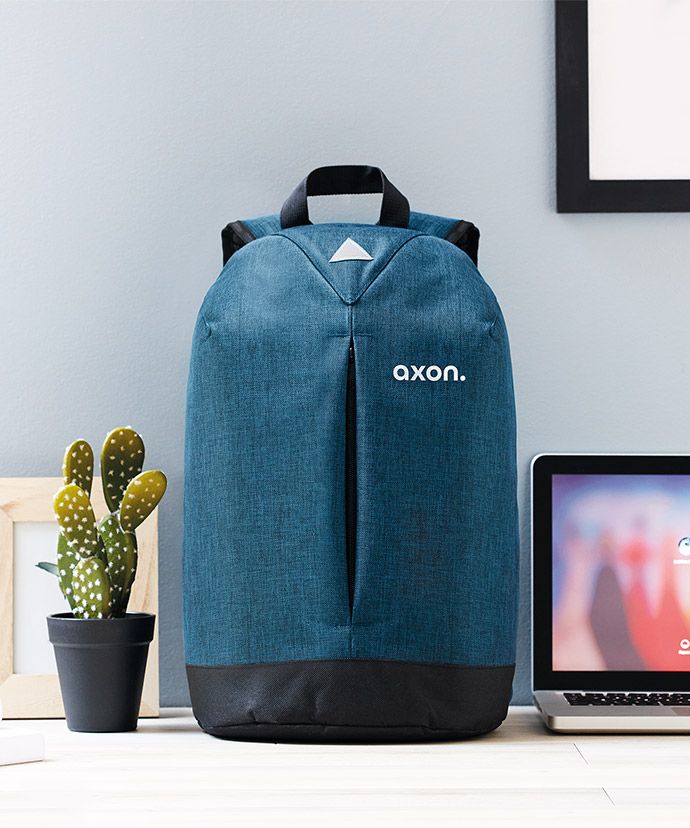 Mochila azul personalizada sobre una mesa con un portátil y una planta. Regalos publicitarios para marketing interno.